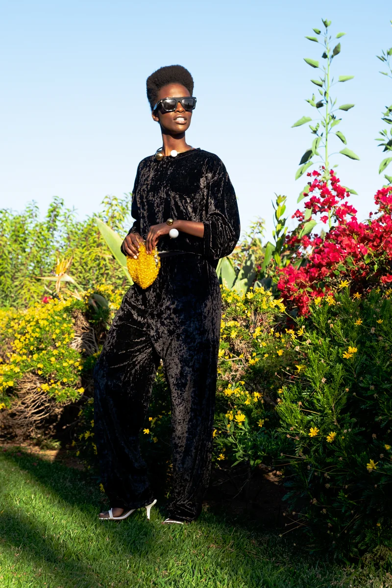 Full-length view of a model in a black velvet outfit and sunglasses holding a yellow bag in a flower garden
