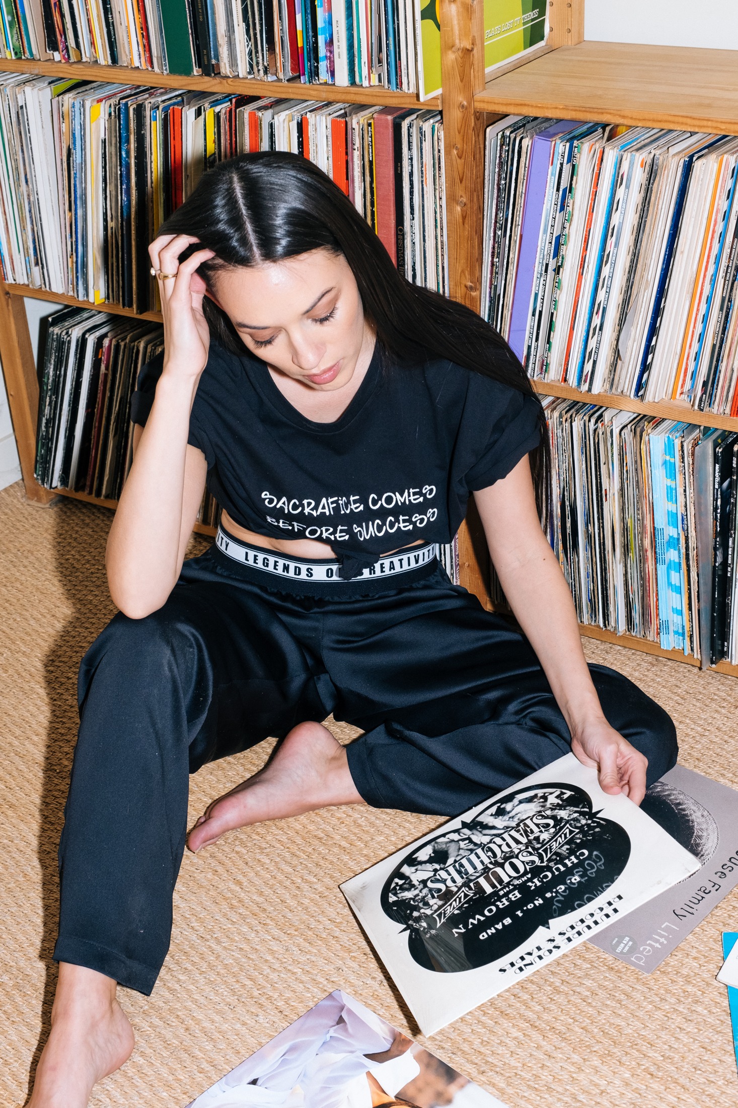 Model seated on the floor browsing records, wearing a black Sacrifice Comes Before Success tee and black satin trousers with the Legends of Creativity waistband