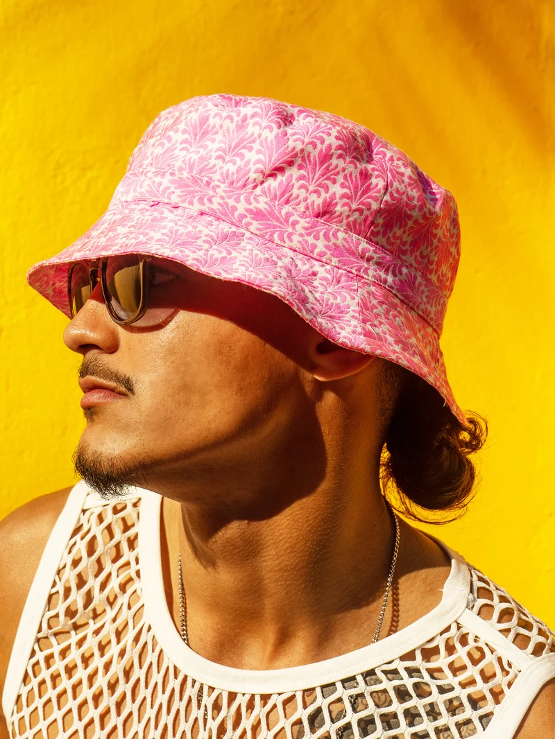 Profile portrait of a model in a pink printed bucket hat, sunglasses and white mesh top against a yellow wall