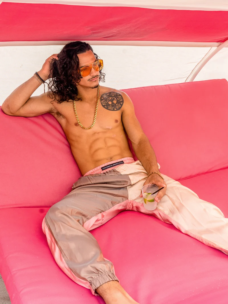 Model reclining on a pink cabana sofa in beige, taupe and pink satin trousers, holding a drink