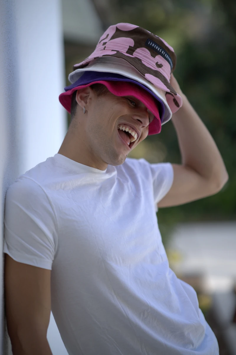 Smiling model in a white T-shirt wearing stacked hot-pink, purple, chalk and pink-and-brown printed bucket hats