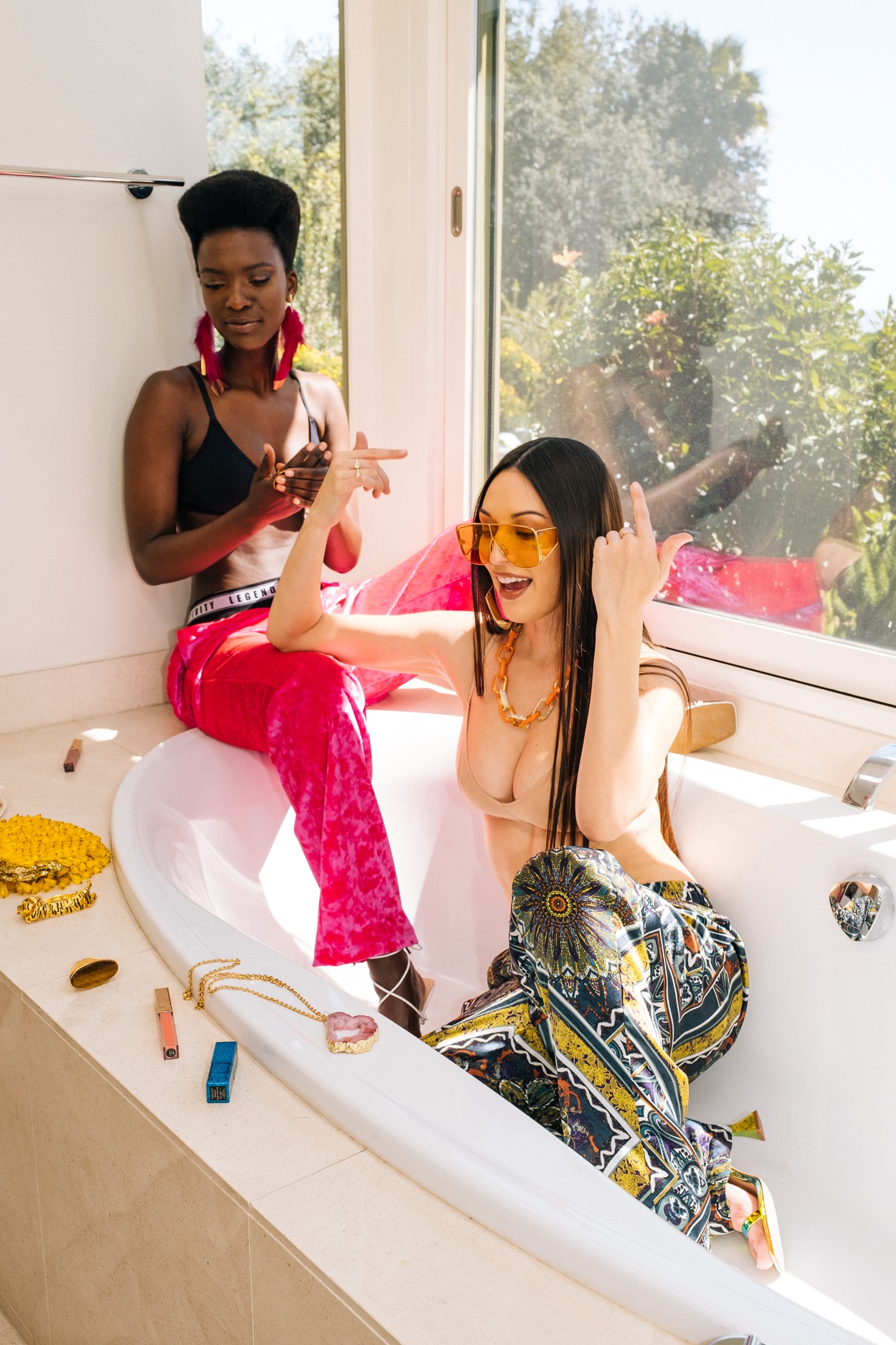 Two models getting ready in a sunlit bathroom, one in hot-pink crushed-velvet trousers with the Legends of Creativity waistband, one in a printed silk set