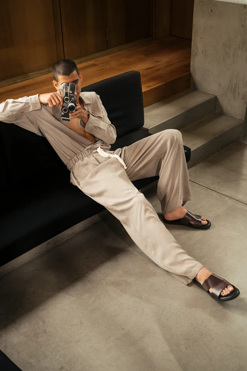 Model in a taupe relaxed shirt and drawstring trousers reclining on a black sofa, holding a vintage camera