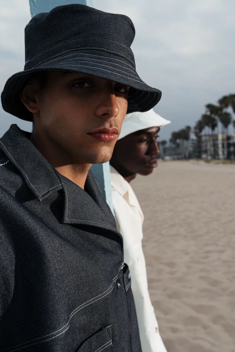 Close portrait at the beach of two models in bucket hats, one in a dark denim jacket with contrast stitching, one in white
