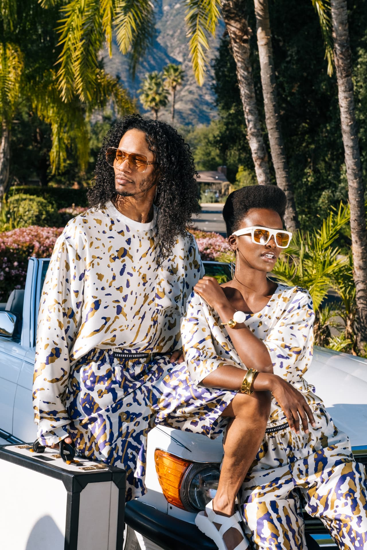 Two models in coordinated white, gold and navy printed silk sets beside a white vintage Mercedes beneath Los Angeles palms