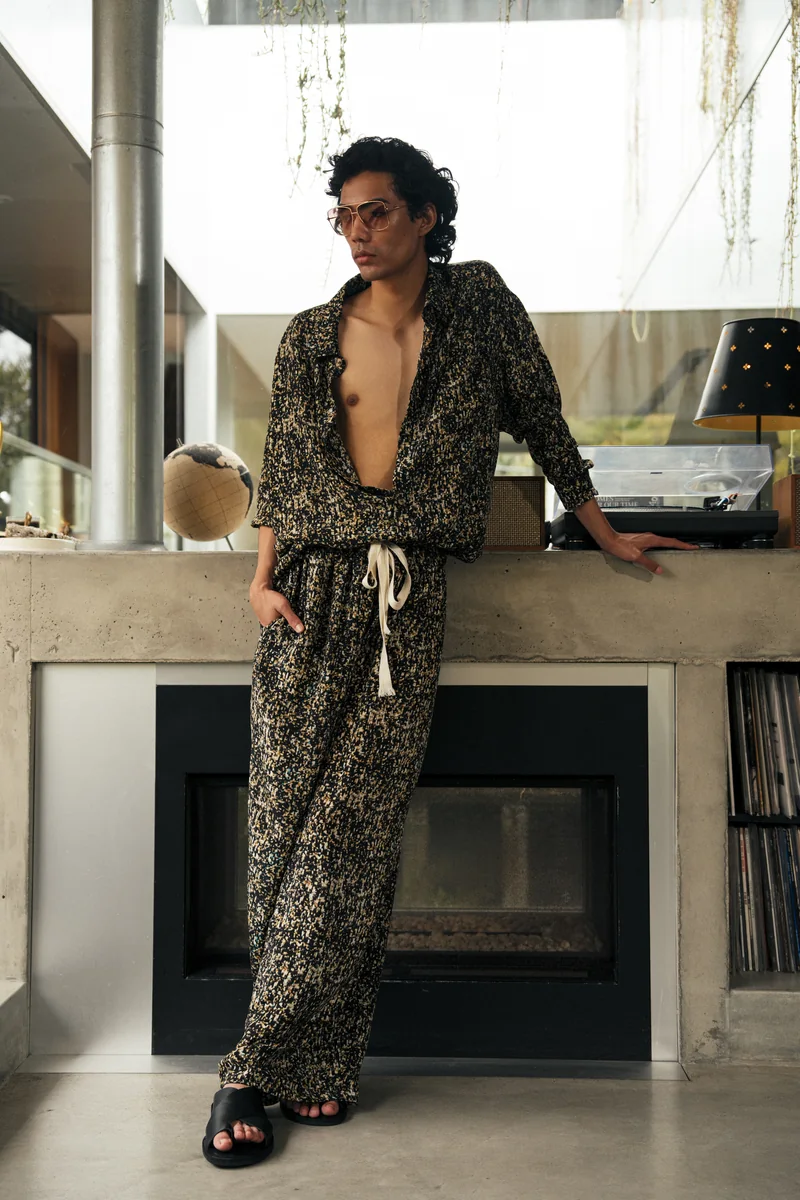 Model in a black and gold printed shirt and trousers leaning against a concrete fireplace