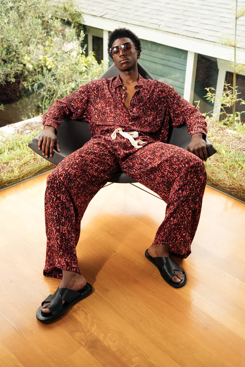 Model in a red and black printed shirt and trousers seated in a black chair on a wooden floor