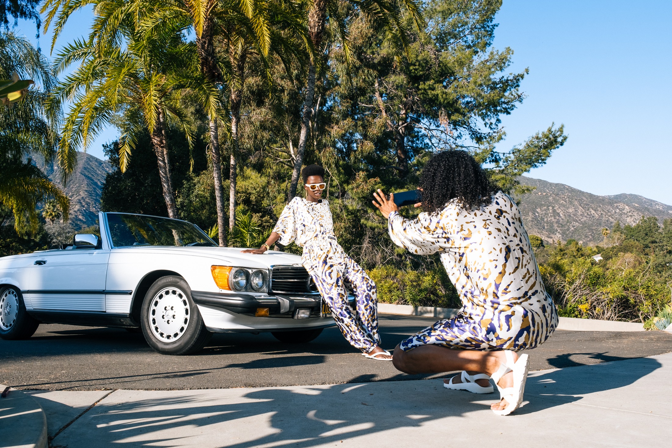 Two models in matching white, gold and violet printed sets photographing each other beside the white Mercedes convertible under palms