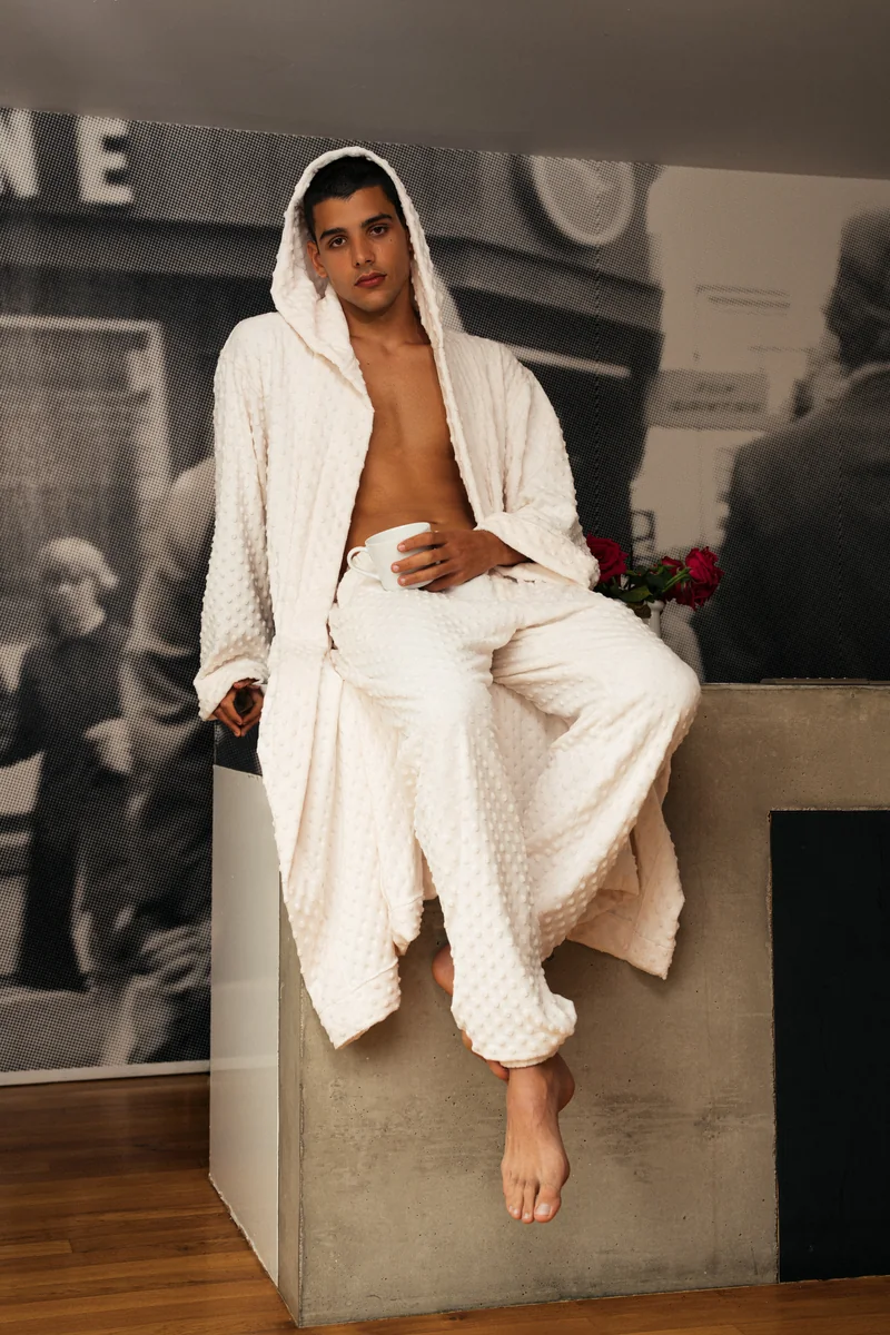 Model in a white textured hooded robe and trousers seated on a concrete ledge, full length