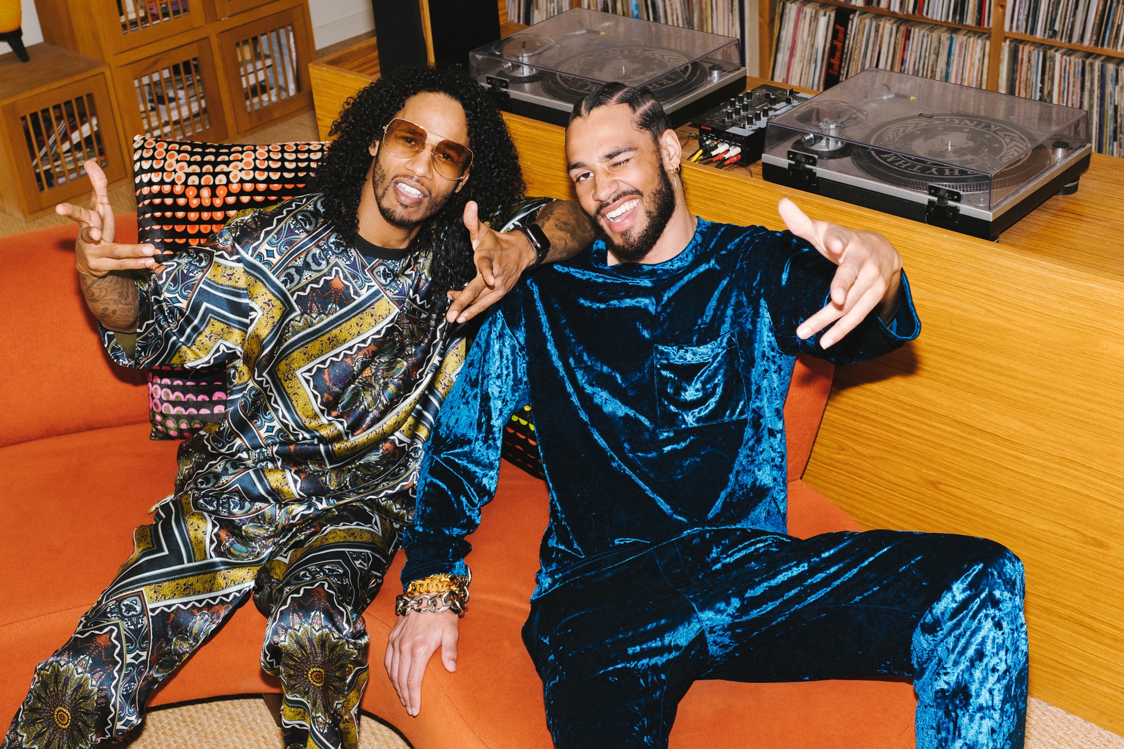 Two models seated on an orange sofa in the record room, one in a printed silk set and one in a teal crushed-velvet set, turntables and record shelves behind