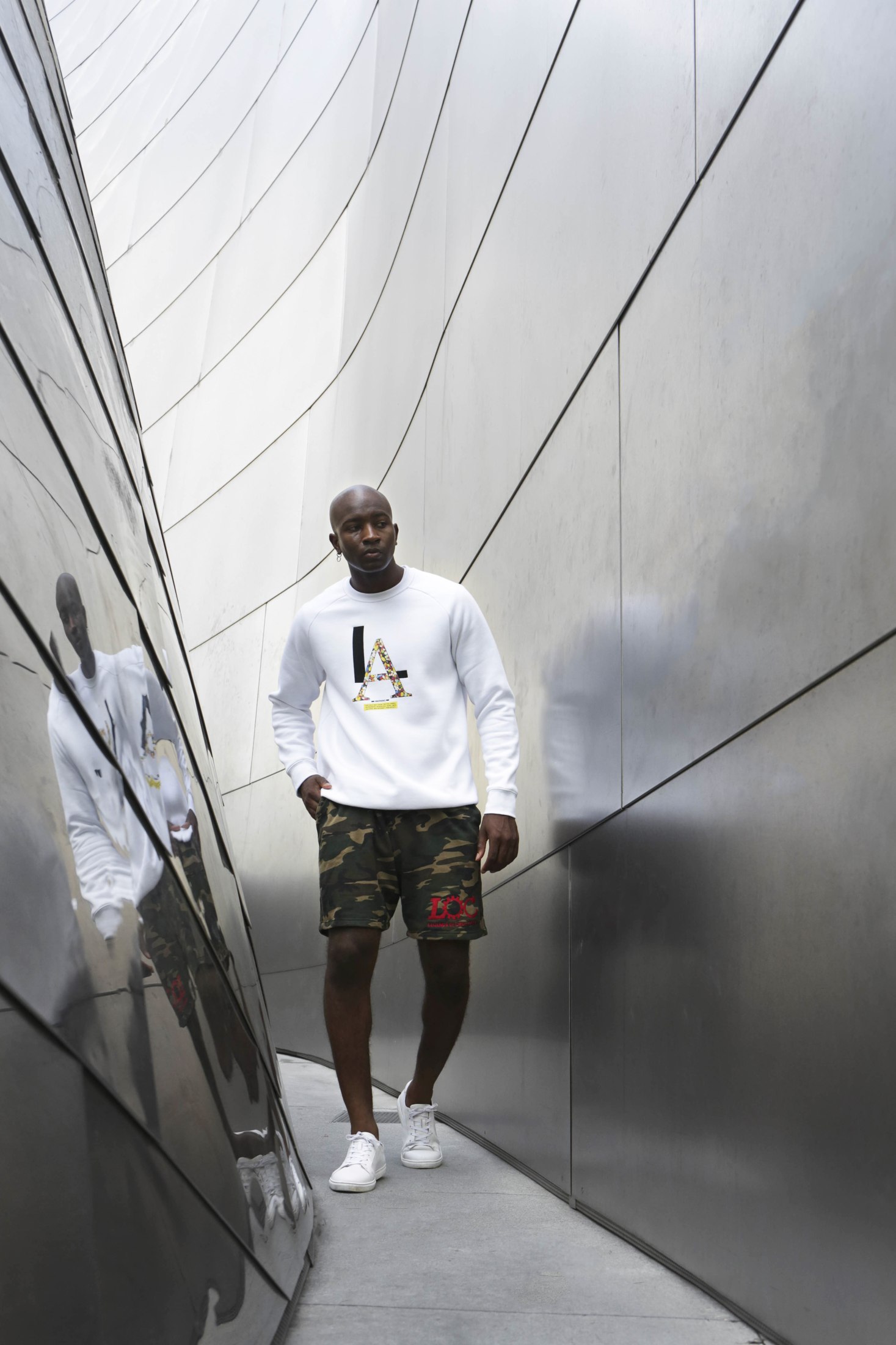 Model in a white graphic sweatshirt and camouflage shorts between metal walls