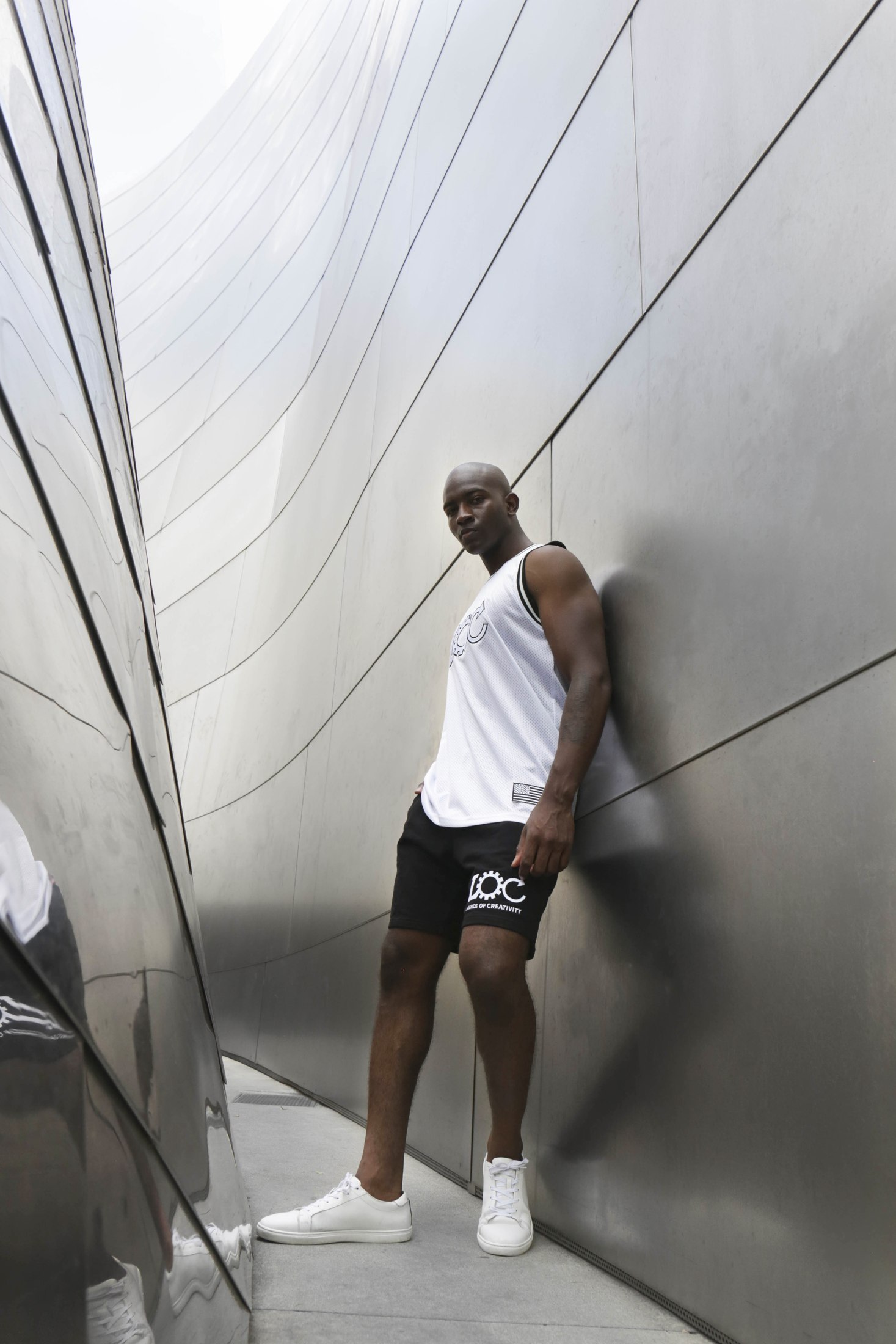 Model in a white sleeveless LOC top and black shorts beside a metal wall
